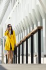 Sorrindo asiático mulher de negócios com casaco amarelo e telefone inteligente andando na rua com edifício no fundo — Fotografia de Stock