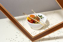 Weiße Schüssel mit leckerem Poke-Gericht und Essstäbchen hinter Gestell auf Tisch mit Sesam bedeckt platziert — Stockfoto