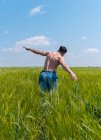 Visão traseira do homem com tronco nu em ganga espalhando braços separados enquanto corria para a frente em grama alta do campo rural — Fotografia de Stock