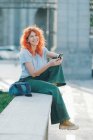 Вид збоку весела руда жінка сидить на вулиці і обмінюється повідомленнями в соціальних мережах на мобільному телефоні, слухаючи музику і дивлячись на камеру — стокове фото