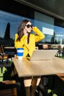 Улыбающаяся азиатская деловая женщина в желтом пальто сидит за столом и пьет кофе со своим смартфоном и ноутбуком — стоковое фото