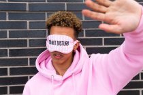 Unrecognizable young curly haired ethnic guy in pink hoodie and blindfold with Do Not Disturb text doing stop gesture against brick wall — Stock Photo