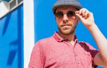 Young bearded guy wearing casual red polo shirt and cap standing against blue wall on street in summer day — Stock Photo