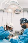 Профессиональный компетентный ветеринарный хирург с ассистентом в защитной одежде и масках, выполняющий операцию на животном пациенте в операционной с хирургической лампой — стоковое фото