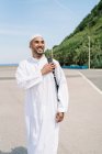 Heureux homme islamique en vêtements traditionnels ajuster sac à dos et regarder loin avec le sourire tout en passant une journée d'été ensoleillée sur la plage — Photo de stock
