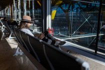 El tipo en el sombrero en el aeropuerto en la sala de espera sentado esperando su vuelo, con auriculares inalámbricos para escuchar música mientras charla con su teléfono inteligente, vista lateral - foto de stock