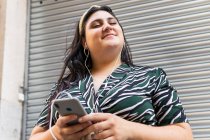 Glückliche junge kurvige Frau in stylischem Zierkleid und Stirnband hört Audio-Nachrichten per Handy mit Kopfhörer und lacht, während sie an der Wand eines modernen Gebäudes steht — Stockfoto