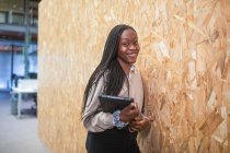Усміхнена афроамериканська жінка-підприємець стоїть з планшетом біля стіни в боягузливому просторі, дивлячись на камеру. — стокове фото