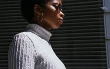Vista lateral de la joven mujer afroamericana en suéter elegante y gafas de sol mirando hacia otro lado mientras está de pie en la luz del sol brillante contra el fondo negro - foto de stock