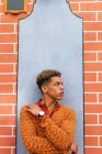 Joven y elegante chico de pelo rizado étnico reflexivo en traje de moda apoyado contra la pared de ladrillo en la calle urbana mirando hacia otro lado - foto de stock