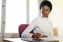 Чорношкіра жінка-лікар, яка пише інформацію на паперовому аркуші під час підготовки медичної доповіді за столом в офісі сучасної клініки — стокове фото