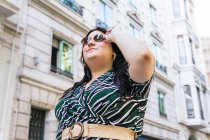 Low angle of confident young curvy female in stylish dress with geometric print and trendy sunglasses looking away while standing near stone urban building in summer day — Stock Photo