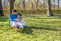 Vista lateral de sonriente pareja de hombres homosexuales sentados en el césped en el parque y disfrutando de un día soleado mientras se miran el uno al otro - foto de stock