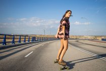 Belle jeune femme patinant longue planche par un pont vide au coucher du soleil — Photo de stock