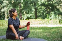 Vue latérale de la jeune femme fatiguée aux cheveux foncés en tenue de sport assise sur un tapis sur une pelouse herbeuse avec des jambes croisées et de l'eau potable après une formation de yoga dans le parc — Photo de stock