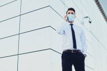 Стильный мужчина в формальной одежде и медицинской маске разговаривает по мобильному телефону, глядя в город — стоковое фото