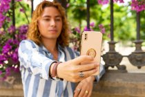Handsome male with long hair and in trendy outfit taking selfie on mobile phone while standing in summer park with blooming trees — Stock Photo