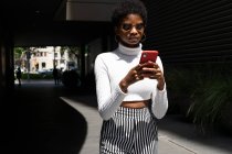 African American woman in trendy clothes browsing social media on cellphone while walking on city street on sunny day — Stock Photo