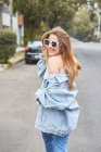 Вид ззаду на стильну молоду жінку в сонцезахисних окулярах і джинсовий одяг сміється на вулиці, насолоджуючись вихідними і дивлячись на камеру над плечем — стокове фото