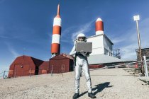 Astronauta macho de cuerpo completo en traje espacial navegando datos en netbook mientras está de pie fuera de la estación con antenas en forma de cohete - foto de stock