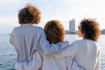 Rückansicht anonymer Freunde mit lockigem Haar, die eng umschlungen vor Stadtbild und Meer im Sonnenlicht stehen — Stockfoto