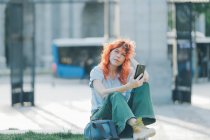 Боковой вид жизнерадостной рыжеволосой женщины, сидящей на улице и сидящей в социальных сетях на мобильном телефоне, слушая музыку и глядя в камеру — стоковое фото