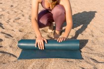 De dessus recadré méconnaissable jeune femme en vêtements de sport placer tapis de yoga sur le sable tout en se préparant pour la pratique sur la plage près de l'océan — Photo de stock