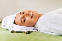 Top view joven hembra feliz con toalla en la cabeza sonriendo y masajeando la cara con rodillo de jade durante la rutina de cuidado de la piel en casa - foto de stock