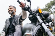 D'en bas motard ethnique masculin barbu en veste en cuir noir assis sur moto moderne sur route asphaltée au milieu d'arbres verts luxuriants poussant dans la vallée montagneuse — Photo de stock