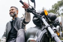 Desde abajo barbudo ciclista masculino étnico en chaqueta de cuero negro sentado en la motocicleta moderna en el camino de asfalto en medio de exuberantes árboles verdes que crecen en el valle montañoso - foto de stock