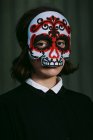 Mysteriöses Weibchen in bemalter Halloween-Maske in Schädelform blickt auf dunklem, verschwommenem Hintergrund in die Kamera — Stockfoto