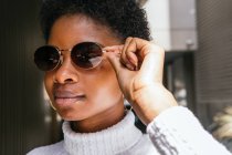 Mujer afroamericana joven en suéter elegante y gafas de sol mirando hacia otro lado mientras está de pie en la luz del sol brillante contra el fondo del edificio de metal - foto de stock
