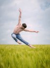 Чоловік без сорочки в денімі, виконуючи чуттєвий балетний стрибок з руками, розкиданими над високою травою в похмурому полі — стокове фото