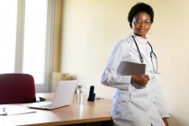 Позитивний молода чорна жінка-лікар в медичному пальто і окулярах зі стетоскопом дивиться на камеру під час роботи з планшетом в сучасному клінічному офісі — стокове фото