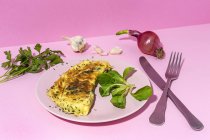 Leckeres Omelett auf Teller gegen frische Petersilienzweige und rote Zwiebel mit Knoblauchzehen auf rosa Hintergrund — Stockfoto