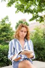 Emotionsloser Mann mit langen Haaren sitzt am Wochenende auf Bank im Garten und liest interessante Geschichte in Buch — Stockfoto