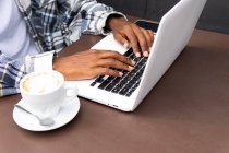 Desde arriba recortado irreconocible afroamericano freelancer masculino navegando y trabajando de forma remota en el ordenador portátil en la cafetería al aire libre mientras está sentado en la mesa con una taza de café - foto de stock