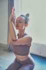 Mujer joven en ropa deportiva realizando pose Garudasana con las manos mientras practica yoga en casa habitación en contraluz - foto de stock