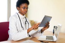 Joven doctora negra en abrigo médico y gafas con estetoscopio trabajando con tableta en consultorio clínico moderno - foto de stock