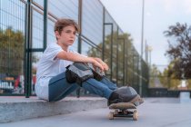 Side view of teen boy sitting with skateboard on ramp in skate park and looking away — Stock Photo