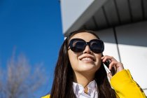 Mujer de negocios asiática sonriente con abrigo amarillo y teléfono inteligente caminando en la calle con el edificio en el fondo - foto de stock