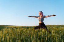 Donna adulta in abiti sportivi praticare yoga con le braccia tese sul prato sotto il cielo blu — Foto stock
