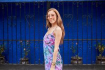 Femme joyeuse portant des lunettes de soleil à la mode et robe d'été debout dans la rue sur fond bleu et regardant la caméra — Photo de stock