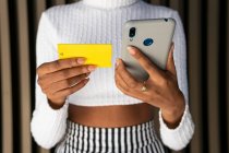 Cortado irreconhecível jovem afro-americano feminino em roupas da moda usando cartão de crédito e smartphone para pagar a ordem on-line contra a parede listrada na rua — Fotografia de Stock