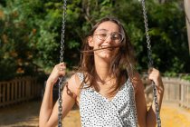 Весела молода жінка в окулярах гойдається в парку в сонячний літній день, дивлячись на камеру — стокове фото