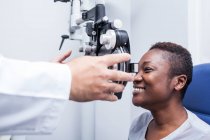 Optometrist adjusting the optometry equipment during study of the eyesight of a happy black woman — Stock Photo