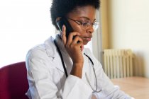 Competente joven afroamericana practicante médica contesta llamada telefónica y el uso de la computadora portátil mientras consulta a los pacientes de forma remota desde el consultorio - foto de stock
