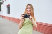 Fotógrafa feminina positiva com câmera tirando foto na rua da cidade enquanto desfruta de fim de semana de verão — Fotografia de Stock