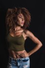 Capelli ricci donna afroamericana in crop top alla moda e jeans in piedi con mano in vita su sfondo nero in studio — Foto stock