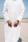 Crop Islamic male in traditional white clothes standing on rug and praying against blue sky on beach — Stock Photo